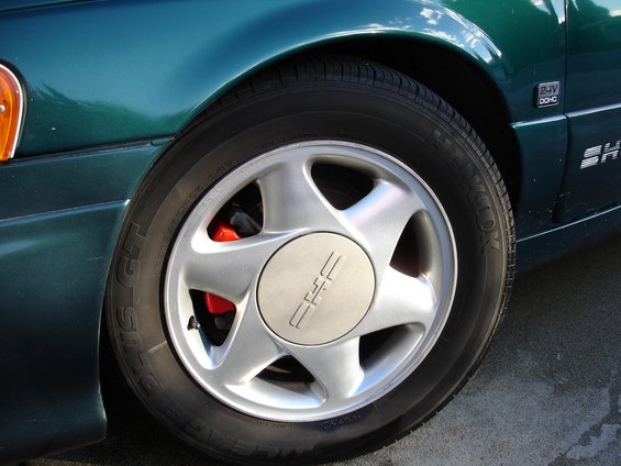 Close-up of a green sedan's front wheel and fender. The wheel has a silver five-spoke design with a center cap showing three vertical lines. Red brake calipers are visible behind the spokes. A '24V DOHC' badge is on the fender