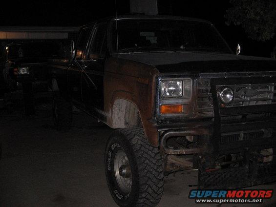 1980s Ford pickup truck with aftermarket bumper and rust on fender. Another truck is visible behind it