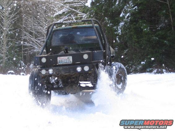 Truck with a roll cage and flatbed driving through snow, kicking up spray
