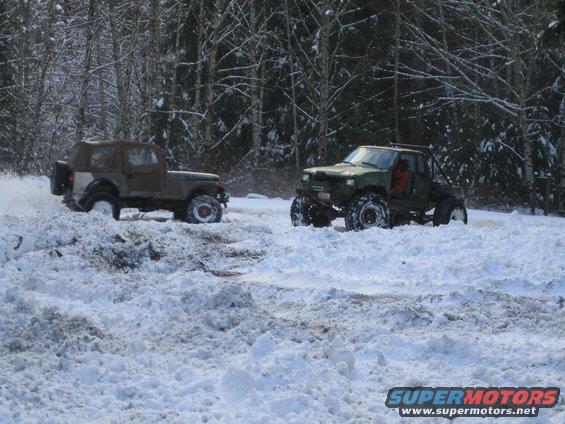 Two modified off-road vehicles driving through deep snow in a wooded area