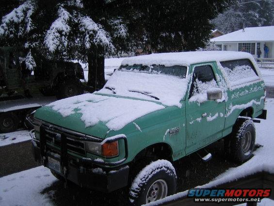 Green 1990 Ford Bronco covered in snow, parked next to another vehicle