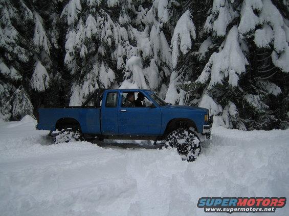 Blue pickup truck driving through deep snow with snow-covered trees in background