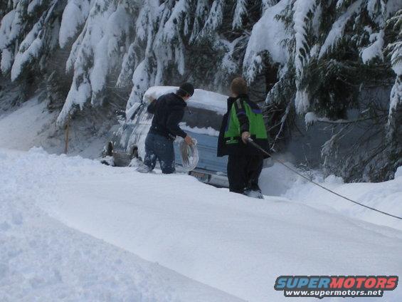 Two people are trying to pull a vehicle stuck in deep snow next to a wooded area