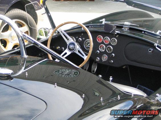 Interior of a dark-colored Shelby Cobra with a wood-rimmed steering wheel and multiple gauges on the dashboard. Another car is visible to the left