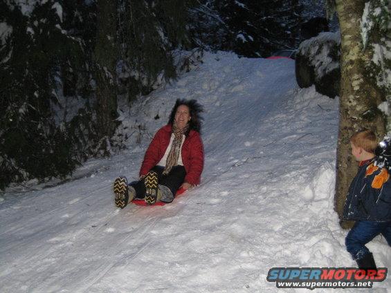 Woman on a red sled going down a snow-covered hill, with a young boy standing to the right