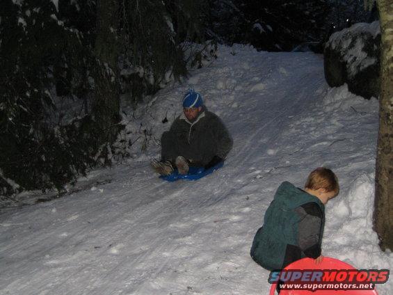 Two people sledding down a snow-covered hill at night, one adult and one child
