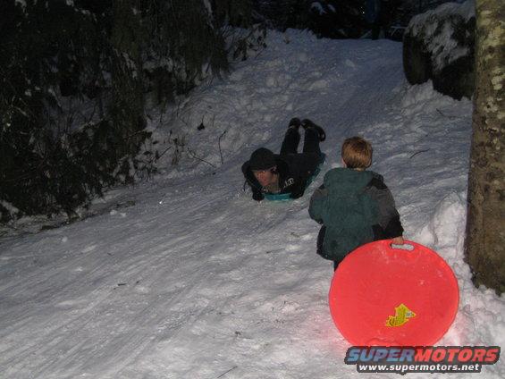Two people sledding down a snowy hill at dusk, one on a blue sled, the other holding a red saucer sled