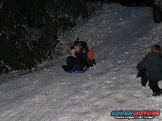 Kid on a blue sled going down a snowy hill at night. Another kid is further down the hill
