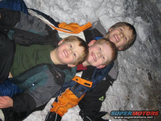 Three smiling boys bundled in snow gear lie together in the snow