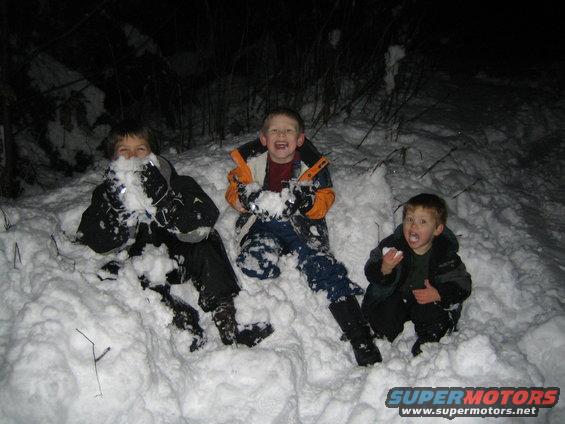 Three boys in snowsuits sit in deep snow, one with snow on his face, another laughing, and the third holding a snowball