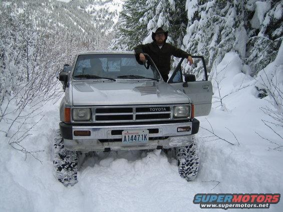 Silver Toyota pickup truck with large tires stuck in deep snow, driver wearing a cowboy hat stands by the open door