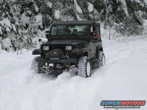 Black Jeep Wrangler driving through deep snow with trees in background