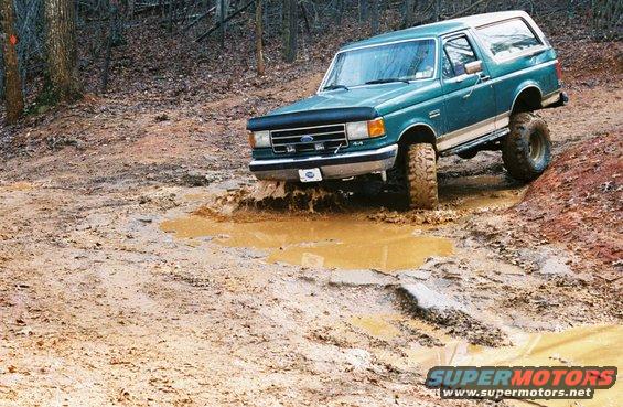 Green 1990 Ford Bronco driving through a muddy puddle on a wooded trail
