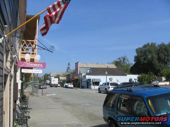 Street view with buildings, an American flag, and several parked and moving vehicles
