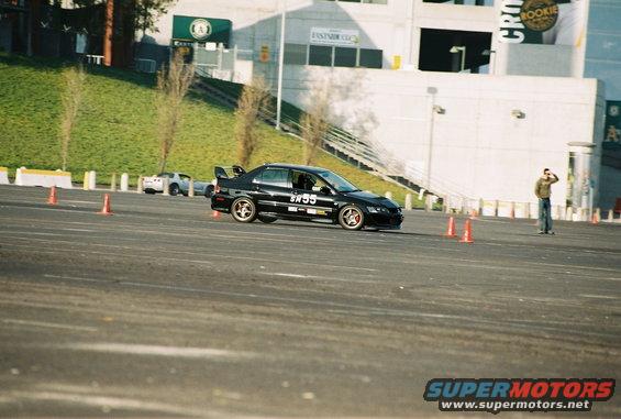 Black Mitsubishi Lancer Evolution with number 55 on the door, driving on a track with orange cones. Another white car is visible in the background