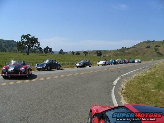Several vintage cars, including a red convertible and a dark coupe, drive in a line on a rural road