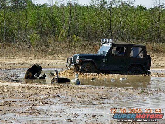Dark green Jeep Wrangler stuck in mud, front bumper visible, soft top up, off-road lights on roof