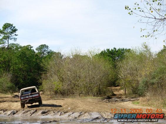 1987 Ford Bronco driving on a muddy off-road trail with trees in the background