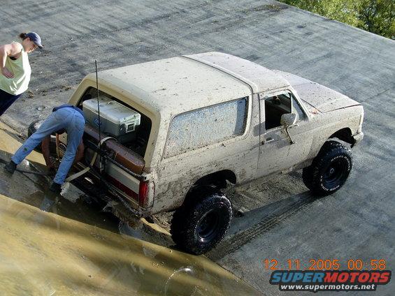 1987 Ford Bronco, 3q-front view, covered in mud. Two people are near the rear