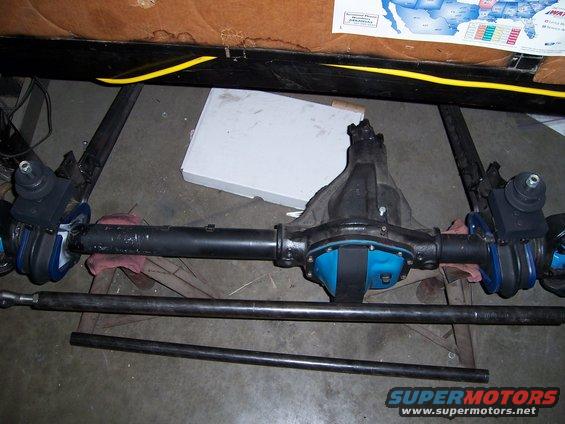 Vehicle rear axle assembly with blue differential cover and steering linkage components. Two metal rods are laid out below it