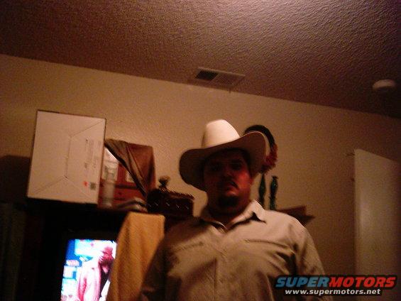 Man wearing a white cowboy hat and a tan shirt stands indoors