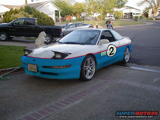 White and blue Ford Probe with racing stripes and a large number 2 on the side. A black pickup truck is parked nearby