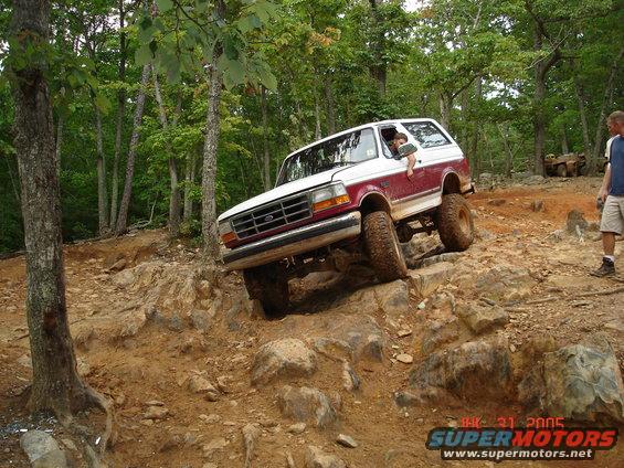 1994 Ford Bronco climbing over large rocks on a wooded trail. Driver visible in window