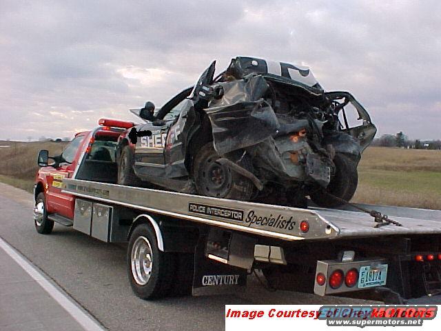 Heavily damaged black police car with 'SHERIFF' visible, loaded onto a red tow truck