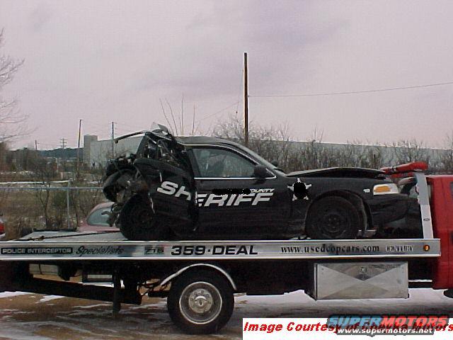 Damaged black sheriff's car with 'SHERIFF' text on its side, loaded onto a tow truck