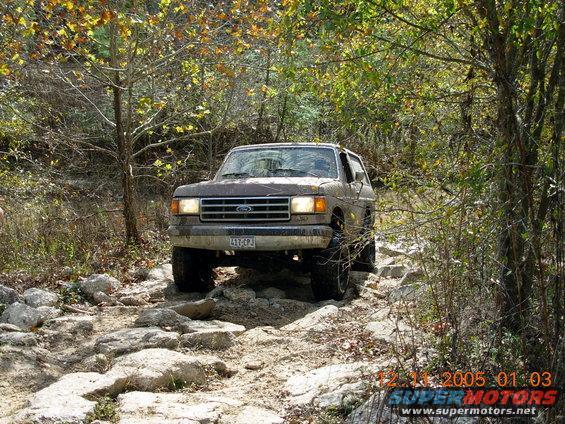 1987 Ford Bronco navigating a rocky off-road path with trees and foliage surrounding it