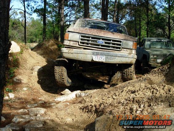 1987 Ford Bronco navigating a muddy, rocky off-road trail with another truck behind it