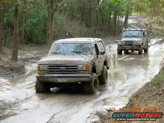 Muddy Ford Bronco and Jeep driving through a wet, muddy trail with trees on the sides