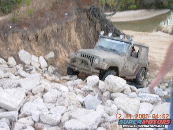 Mud-covered Jeep Wrangler navigating a rocky, eroded embankment next to a body of water