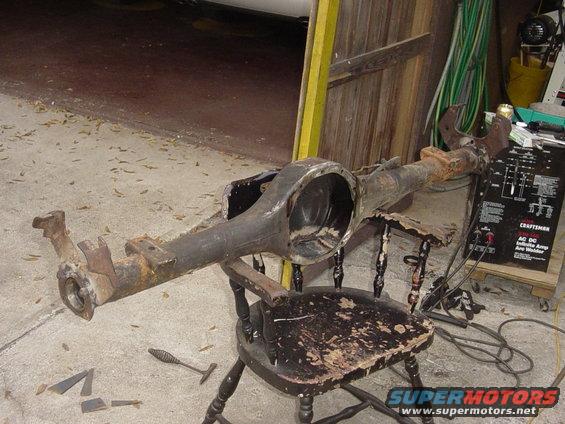 Rusty rear axle assembly resting on a wooden chair next to a welding machine