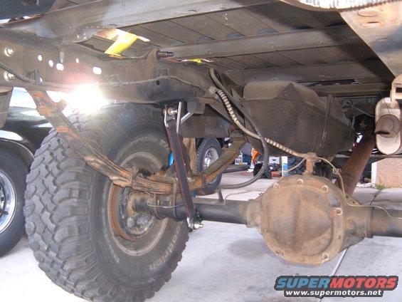 Underside view of a truck's rear axle, leaf springs, and fuel tank. Another vehicle is partially visible on the left