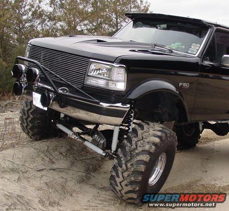 Black 1993 Ford F-150 pickup truck with large off-road tires and a chrome bumper with auxiliary lights, viewed from the front three-quarter angle