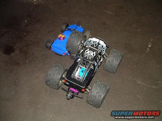 Two remote control monster trucks, one black with flames and one blue, on a dark surface