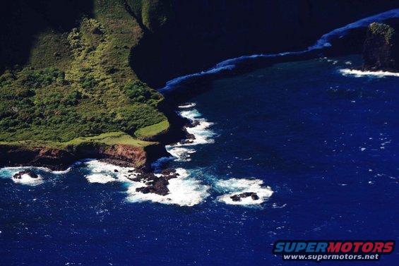 Ocean waves crash against rocky cliffs and a green, vegetated coastline