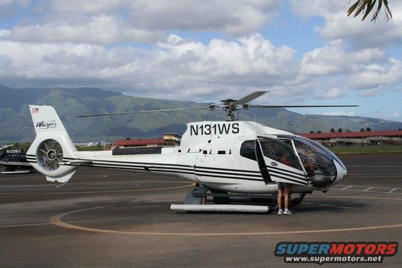 White helicopter with black stripes, registration N131WS, parked on tarmac with mountains in background. Another helicopter visible to the left