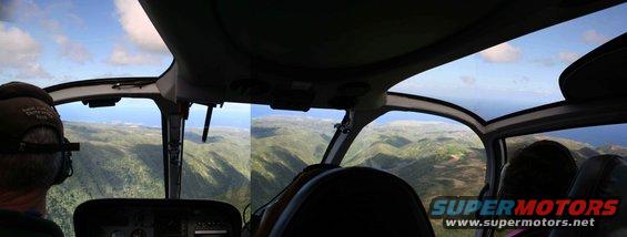View from inside a helicopter cockpit looking out over green mountains and the ocean