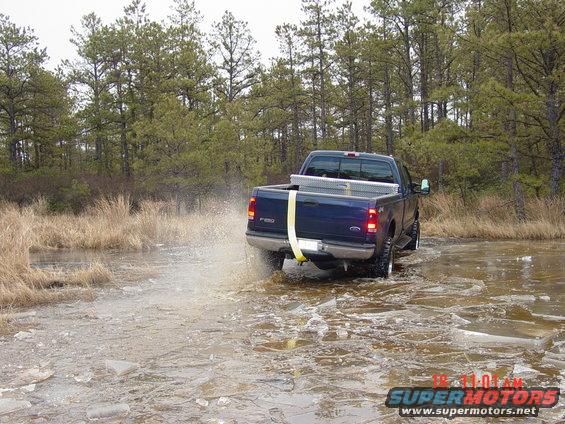 Blue pickup truck with a toolbox in the bed drives through shallow water, kicking up spray. The truck has "F-250" badging