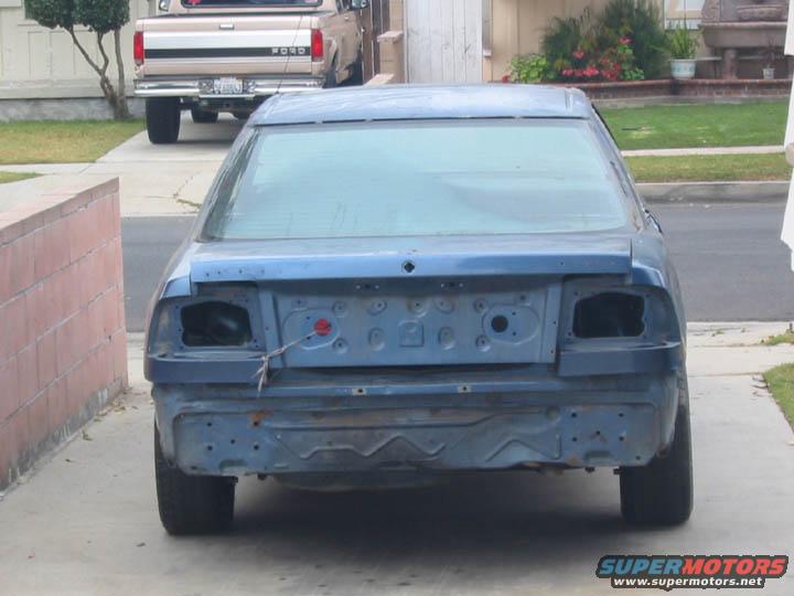 Rear view of a blue car with no bumper or taillights. A Ford pickup truck is visible in the background