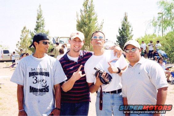 Four guys posing for a photo outdoors, with trees and other people in the background. One guy wears a shirt that says '3rd Annual Mountain 3 on 3 Madness'