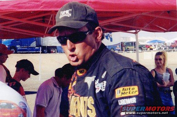 Man in a black cap and racing jacket with patches, sunglasses, and an open mouth. A blue truck is visible in the background