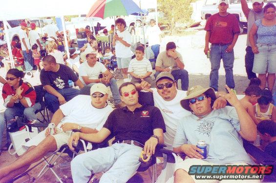 Group of men and women sitting in chairs at an outdoor gathering, with tents and vehicles in the background