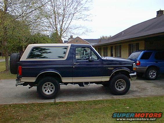 Dark blue Ford Bronco with tan trim and a spare tire on the back, parked next to a blue SUV in a driveway