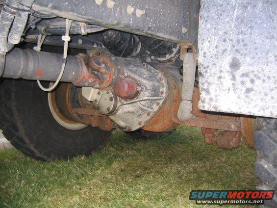 Underside view of a truck's rear axle, driveshaft, and suspension components. Shows a large tire and rusty metal parts
