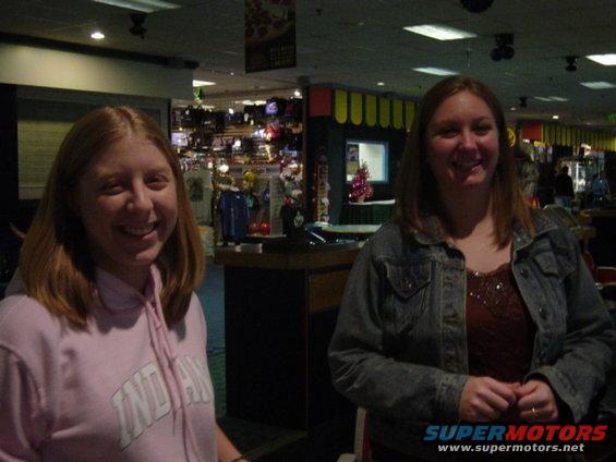 Two women smiling indoors, one in a pink hoodie, the other in a denim jacket