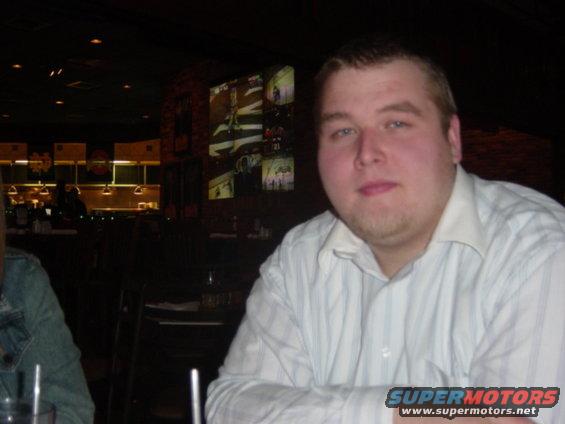 Man in a striped shirt sits at a table in a dimly lit restaurant with screens on the wall behind him