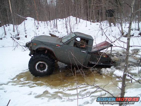 Off-road pickup truck stuck in icy water with trees and snow in background